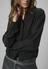 Kobieta z jasnobrązowymi włosami, ubrana w luźną, czarną bluzkę z długim rękawem i szare aksamitne spodnie, trzymająca dłoń na szyi, na jednolitym tle.