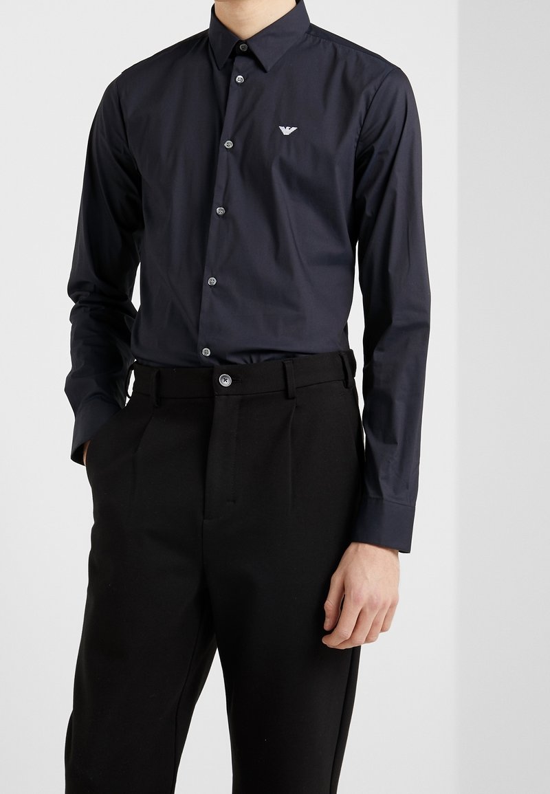 Man som bär en åtsittande svart skjorta med knappar och en liten vit örnlogotyp samt svarta skräddarsydda byxor med händerna i fickorna.