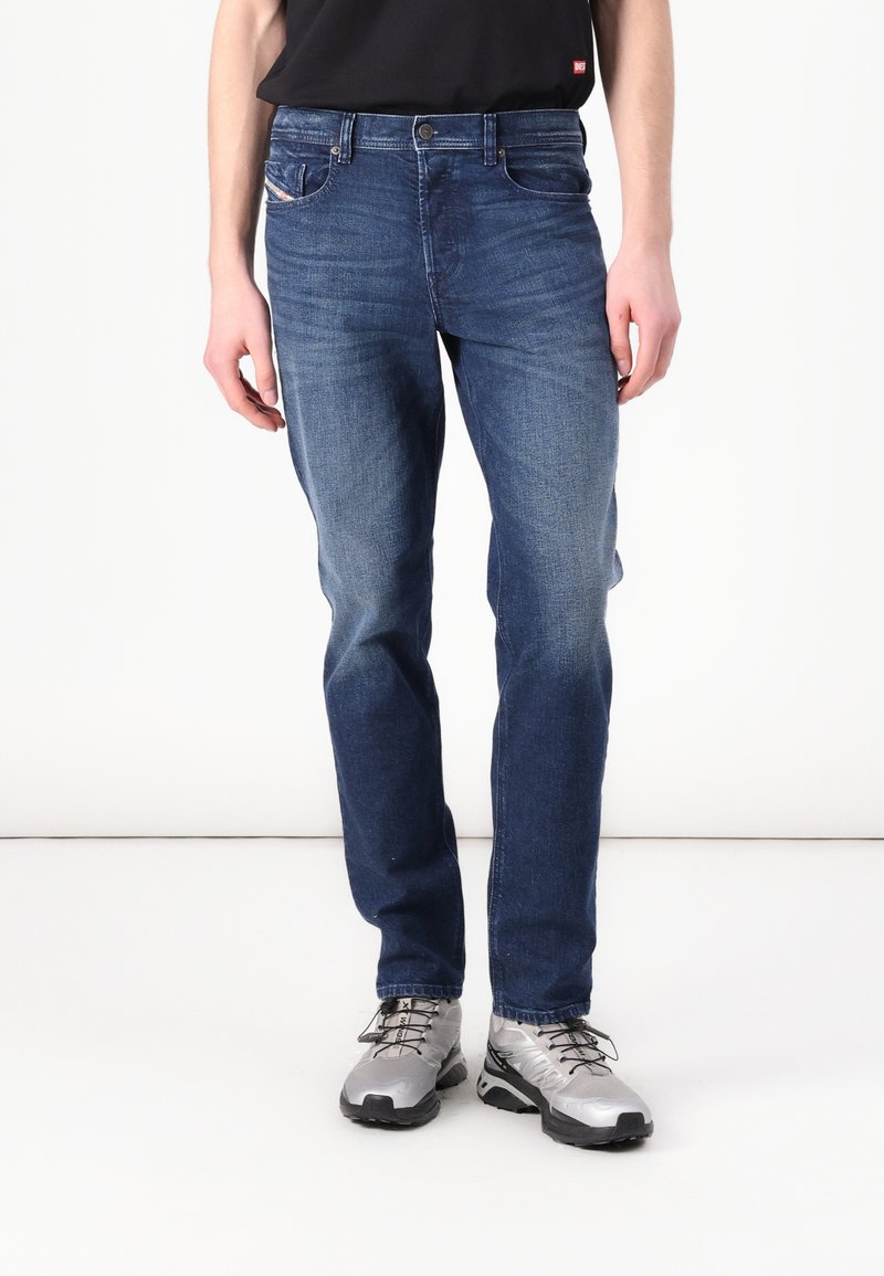 Jean en denim bleu foncé avec une coupe slim, des détails décolorés, un design à cinq poches et une fermeture à bouton argenté. Porté avec des chaussures de sport grises.