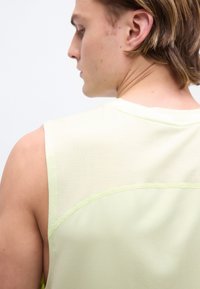 Jeune homme aux cheveux bruns courts portant un débardeur de sport jaune pâle, tourné de profil sur un fond blanc uni.