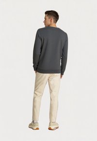 Nuori mies seisoo selkä Camera kohti, päällään pitkähihaisi tummanharmaa villapaita, vaaleat beige housut, valkoiset sukat ja beige urheilukengät.
