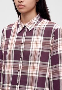 Γυναίκα που φοράει μωβ, λευκό και ανοιχτό καφέ καρό πουκάμισο με γιακά και κουμπιά, εμφανίζεται από τους ώμους μέχρι τα χείλη με γκρι φόντο.