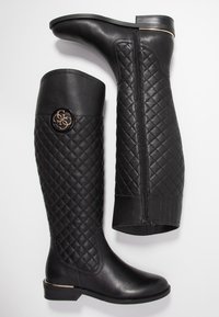 Mustat polvipituiset saappaat, jotka on valmistettu teksturoidsesta nahasta ja joissa on tikattu kuvio, kultasävyinen yksityiskohta ja sivuvetoketju helppoa pukemista varten.