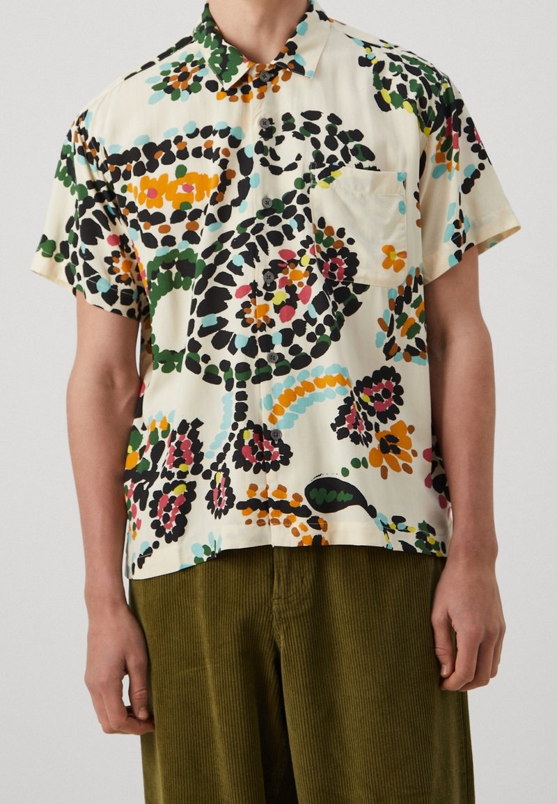 Chemise à manches courtes boutonnée avec un motif floral abstrait coloré portée avec un pantalon en velours côtelé vert olive.