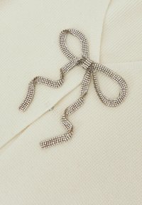 Bílý pletený oděv s jemnou stříbrnou mašlí a třpytivými, perličkovými detaily, které zvyšují celkovou texturu a design.