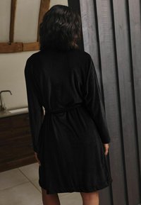Schwarzer, langärmeliger Bademantel mit gebundenem Bund, aus strukturiertem Stoff, der bis zur Mitte des Oberschenkels hängt, von hinten gegen einen neutralen Hintergrund gezeigt.