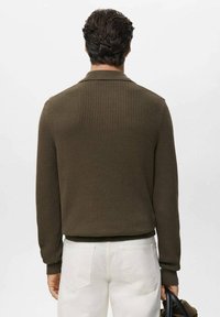 Jersey de canalé verde oliva con cuello y puños ajustados, combinado con pantalones blancos. El diseño presenta una tela texturada y una silueta estilizada.