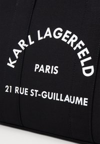 Czarny teksturowany materiał z białym nadrukiem: "KARL LAGERFELD," "PARIS" oraz "21 RUE ST-GUILLAUME," zawierający pogrubioną, nowoczesną typografię.