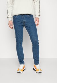 Jeans skinny azuis feitos de denim, com cinco bolsos e um fecho clássico com botão. Usados com um hoodie de cor clara e ténis desportivos.