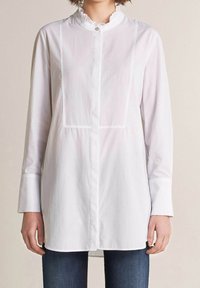 Biała koszula z długim rękawem i falbaną przy dekolcie, z pionowymi przeszyciami. Wykonana z lekkiej bawełnianej tkaniny.