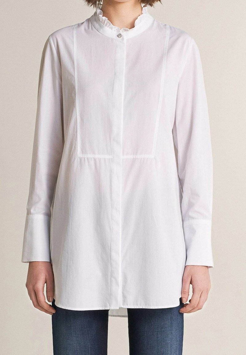 Biała koszula z długim rękawem i falbaną przy dekolcie, z pionowymi przeszyciami. Wykonana z lekkiej bawełnianej tkaniny.