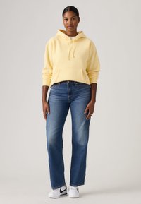 Κίτρινο φούτερ με κουκούλα, μπροστινή τσέπη και κορδόνια, σε συνδυασμό με τζιν μεσαίου μπλε χρώματος και ίσια μπατζάκια, καθώς και λευκά αθλητικά παπούτσια με μαύρες λεπτομέρειες.