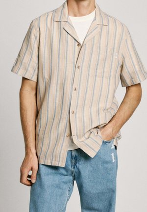 Mann trägt ein beige-blau gestreiftes Kurzarmhemd mit Knöpfen über einem weißen T-Shirt und hellblauen, zerrissenen Jeans.