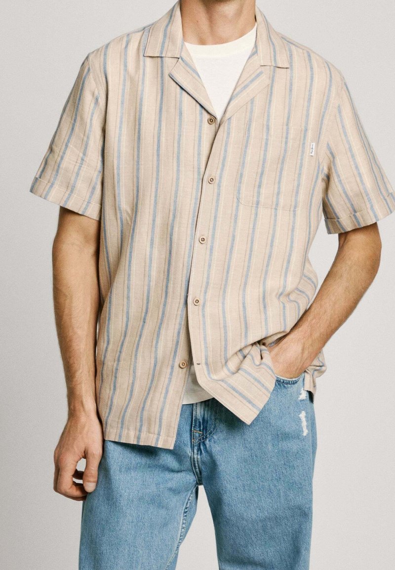 Homme portant une chemise à manches courtes boutonnée à rayures verticales beige et bleu par-dessus un T-shirt blanc avec un jean bleu clair déchiré.