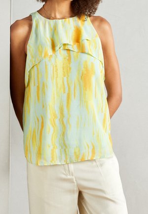 Sleeveless blouse with yellow and light blue abstract pattern, featuring layered ruffle detail at the chest, worn with cream pants.