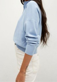 Suéter de punto azul claro con cuello alto, puños acanalados y ajuste holgado. Combinado con pantalones blancos de cintura alta, mostrando una textura suave.