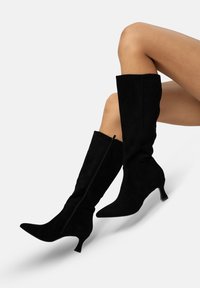 Botas de ante negras hasta la rodilla con punta afilada, tacón estrecho y cremallera lateral. La textura suave y el diseño ajustado realzan la silueta.