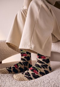 Chaussettes duveteuses et colorées avec un motif léopard, arborant des teintes de noir, rose et vert, portées avec un pantalon de couleur crème.