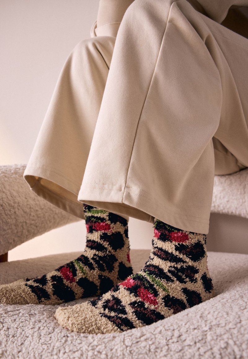 Chaussettes duveteuses et colorées avec un motif léopard, arborant des teintes de noir, rose et vert, portées avec un pantalon de couleur crème.