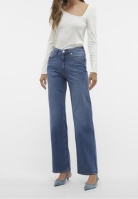 Weißes Langarmshirt mit asymmetrischem Ausschnitt, kombiniert mit hochgeschnittenen, geraden blauen Jeans und hellblauen High Heels.