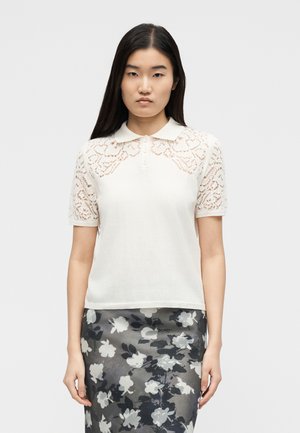 Femeie purtând un top alb cu mâneci scurte din dantelă și guler, și o fustă gri cu model floral, stând pe un fundal simplu.