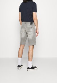 Man som bär blekta grå denimshorts, svarta sneakers, vita strumpor och en mörkblå t-shirt, står vänd bort från kameran mot en neutral bakgrund.