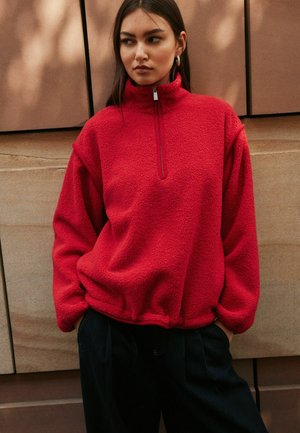 Kvinna med långt mörkt hår som bär en klar röd fleecepullover med halvviss dragkedja och svarta byxor, stående framför en stenvägg.