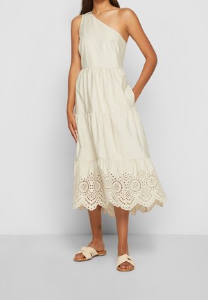 Frau trägt ein cremefarbenes Midi-Kleid mit einem Schulterriemen, Saum aus Lochspitze und beige flache Sandalen mit gedrehten Riemen.
