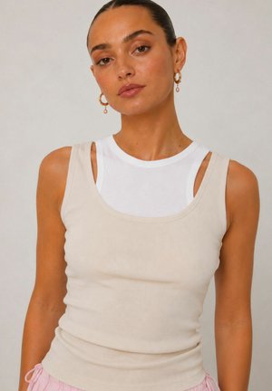 Vrouw met naar achteren gekamd haar draagt gouden oorringen, een witte mouwloze top onder een beige geribde tanktop en lichtroze broek.