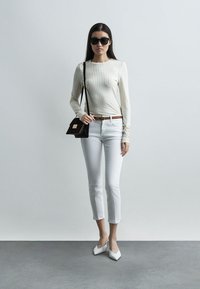 Γυναίκα που φοράει μαύρα γυαλιά ηλίου, κρεμ ριγωτή μπλούζα με μακριά μανίκια, λευκό cropped παντελόνι, λευκά αιχμηρά παπούτσια και μια σκουρόχρωμη τσάντα ώμου.