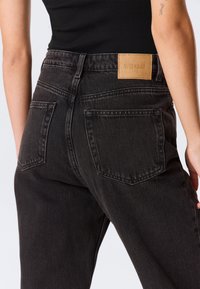 Personne portant un jean noir taille haute avec des poches arrière et un patch beige de la marque Weekday sur la ceinture, associé à un haut noir.