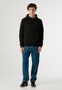 Μαύρη φούτερ μπλούζα με κουκούλα, μπροστινές τσέπες, ρυθμιζόμενο κορδόνι και μικρό λογότυπο. Συνδυάζεται με φαρδιά μπλε τζιν παντελόνια και μαύρα αθλητικά παπούτσια.