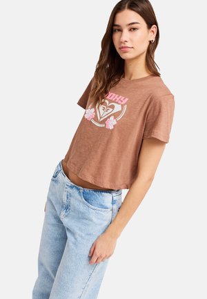 Mladá žena s dlhými hnedými vlasmi, oblečená v hnedom krátkom tričku s ružovými kvetmi a svetlomodrých rifle s vysokým pásom, pózujúca zboku.