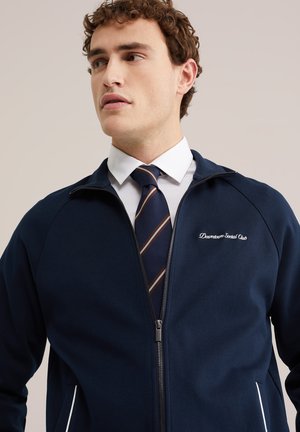 Nooruk lokkis juustega mees kannab laheda sinise tõmblukuga jopet valge särke ja triibulist sinist lipsu taustal.