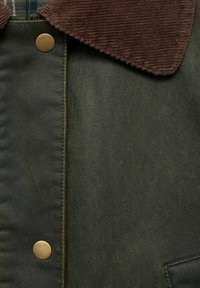 Vihreä kangastakki, jossa on ruskea corduroy-kaulus, metallinapit ja teksturoitu kangas, jossa on näkyvä taskuyksityiskohta.