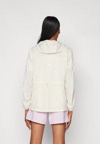 Leichte, creamfarbene Jacke mit Kapuze, elastischen Bündchen und verstellbarem Bund. Verfügt über ein reflektierendes Detail auf dem Rücken.