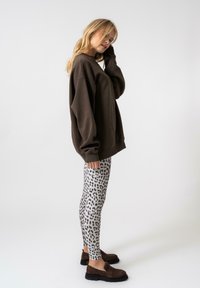 Barna, oversized kapucnis pulóver fehér leggingsszel párosítva, fekete leopárdmintával. A modellen sötét barna slip-on cipő van.