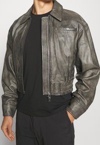 Man med en oöppen grå läderbombarjacka över en svart skjorta och svarta byxor, står med en hand i fickan.