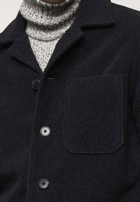 Chaqueta de camisa oscura de mezcla de lana con cuello, que incluye un bolsillo en el pecho y botones redondeados. Suéter de punto texturizado y cálido debajo.