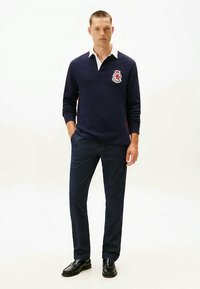 Mężczyzna ubrany w granatową koszulkę rugby z białym kołnierzykiem, ciemne spodnie i czarne buty, stojący z jedną ręką w kieszeni na białym tle.
