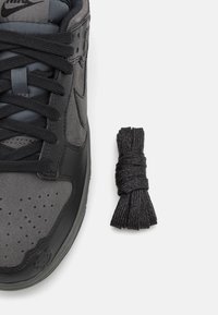 Baskets grises et noires avec des lacets noirs noués, à côté d'une paire supplémentaire de lacets noirs sur un fond blanc.