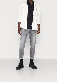 Weiße Jacke mit Knöpfen über einem schwarzen Hemd, kombiniert mit zerrissenen grauen Jeans und schwarzen Schnürboots. Das Outfit zeichnet sich durch ein lässiges, geschichtetes Design aus.