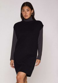 Vestito nero senza maniche in maglia con collo alto, indossato sopra una maglia grigia a maniche lunghe. Presenta un orlo a costine, una vestibilità comoda e spacchi laterali.