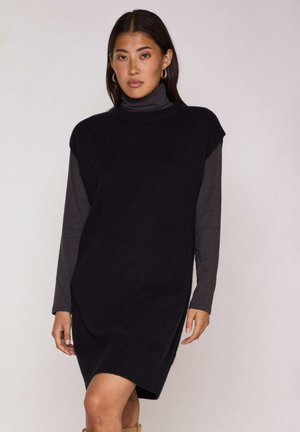 Zwarte gebreide mouwloze jurk met een hoge hals, gedragen over een grijze top met lange mouwen. Voorzien van een geribbelde zoom, een losse pasvorm en zijslitten.