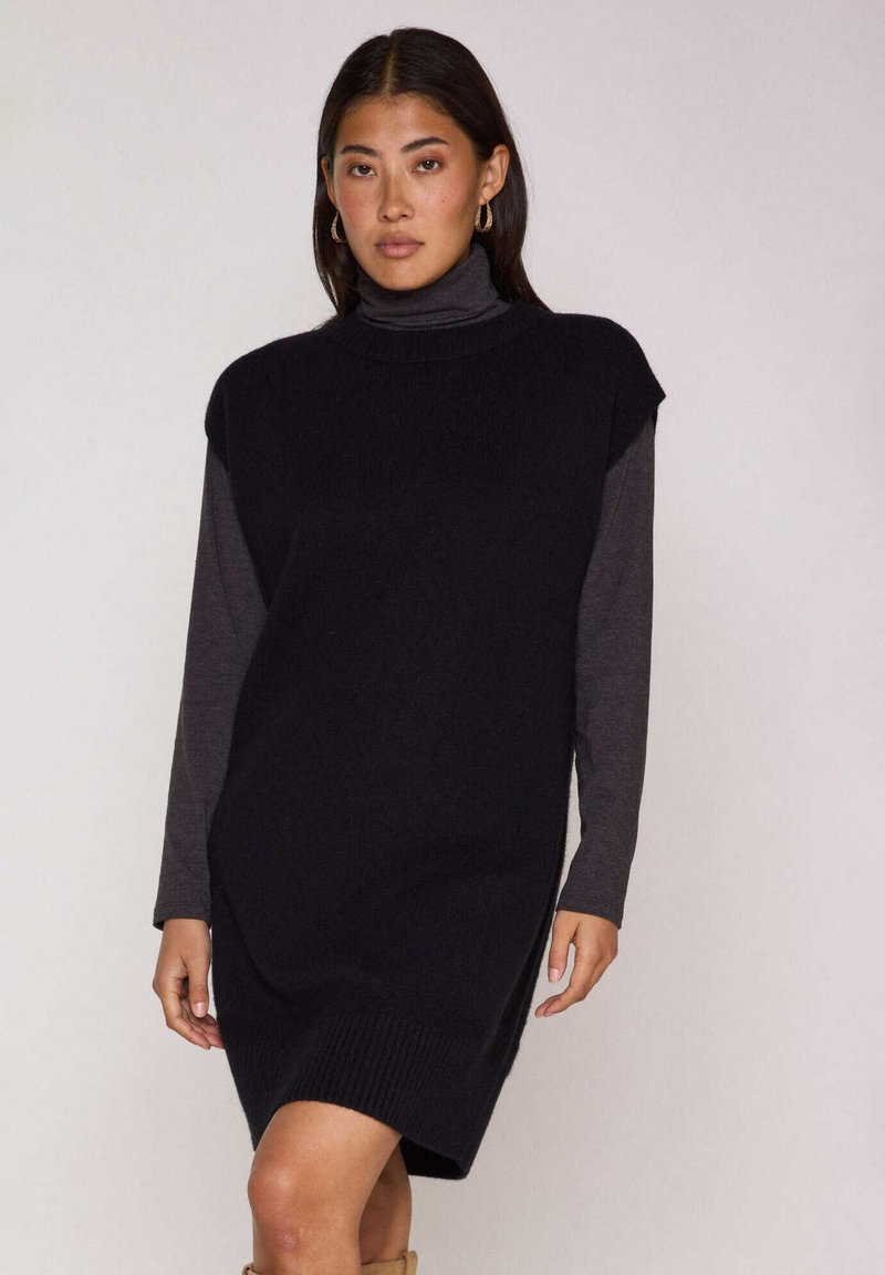 Vestito nero senza maniche in maglia con collo alto, indossato sopra una maglia grigia a maniche lunghe. Presenta un orlo a costine, una vestibilità comoda e spacchi laterali.