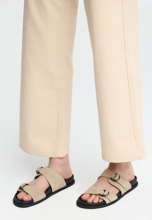 Füße, die beige Wildleder-Sandalen mit breiten Riemen und Metallverschlüssen tragen, kombiniert mit hellfarbenen Hosen mit geradem Bein.