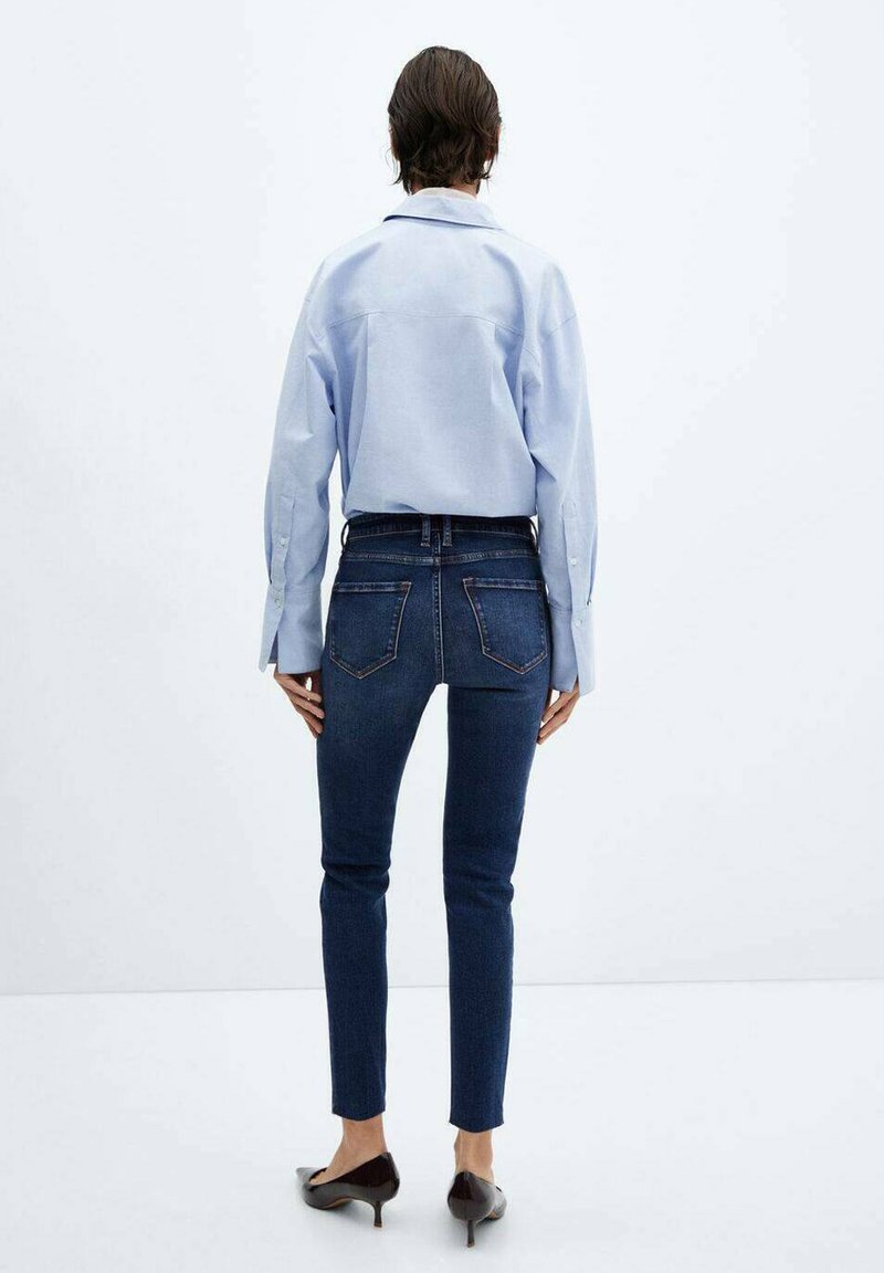 Chemise boutonnée bleu clair à manches longues, associée à un jean bleu foncé ajusté et des chaussures noires à bout pointu, vue de dos.