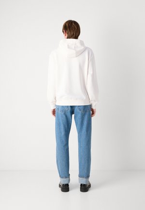 Person trägt einen weißen Hoodie, blaue hochgekrempelte Jeans und schwarze Schuhe und steht mit dem Gesicht zu einem einfarbigen weißen Hintergrund.