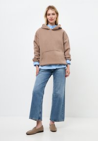 Hanorac oversized de culoare maro-deschis, cu buzunar frontal, asortat cu o cămașă albastră deschisă, blugi largi răniți și pantofi slip-on bej.