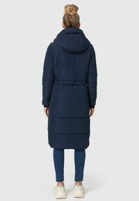 Marineblauwe gewatteerde jas met capuchon en tailleband, voorzien van horizontale naadpatronen en een middellange ontwerp, gedragen over jeans en sneakers.